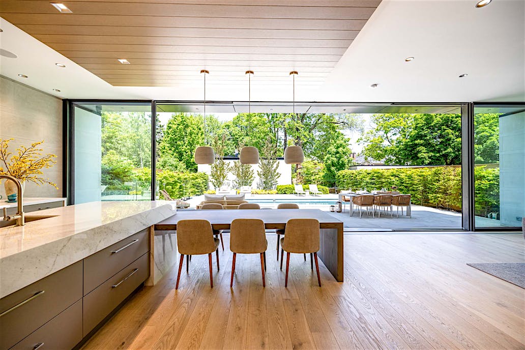 Large sliding glass doors connect a modern kitchen and dining area to a backyard patio with pool, outdoor dining, and lush greenery