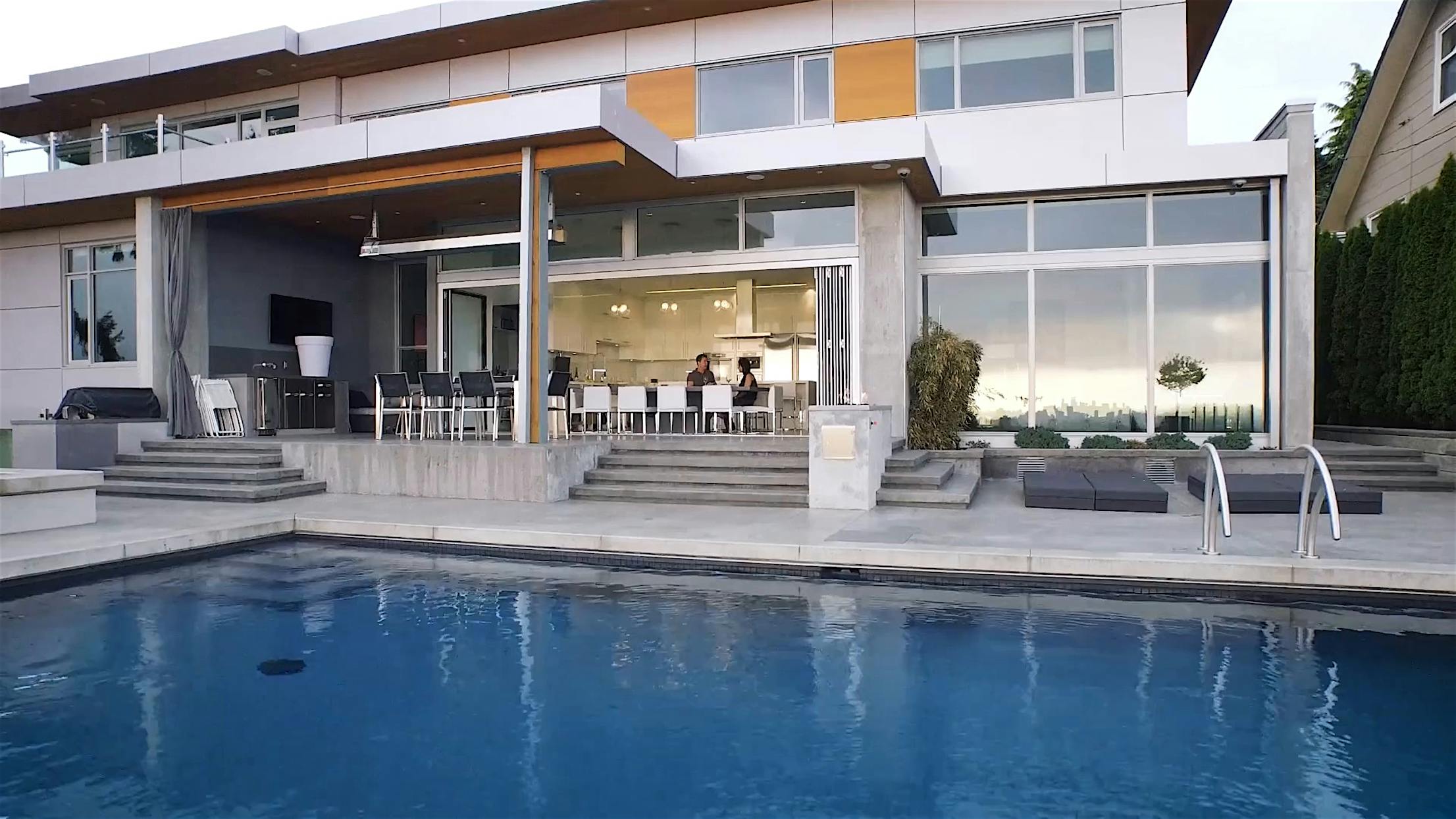 Modern house with large windows, sleek glass walls, an outdoor dining area, and a swimming pool in the foreground.